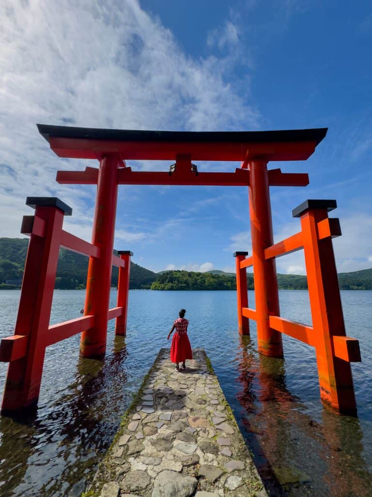 A large red torii gate standing in Lake Biwa with a woman in a red dress standing on the stone path beneath it, surrounded by scenic mountains and a blue sky.