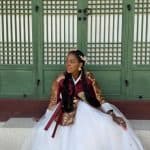 Nma Okafor dressed in traditional Korean hanbok sitting in front of a green wooden door.