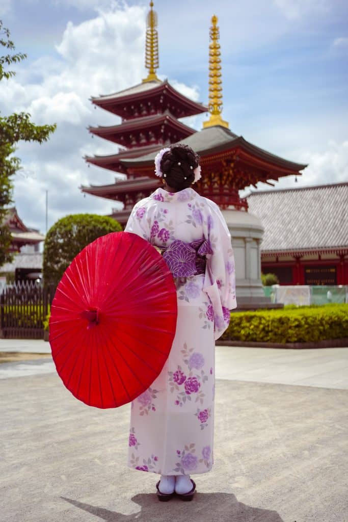 Woman in a floral kimono holding a red parasol at a historic Japanese temple with pagoda architecture in the background.